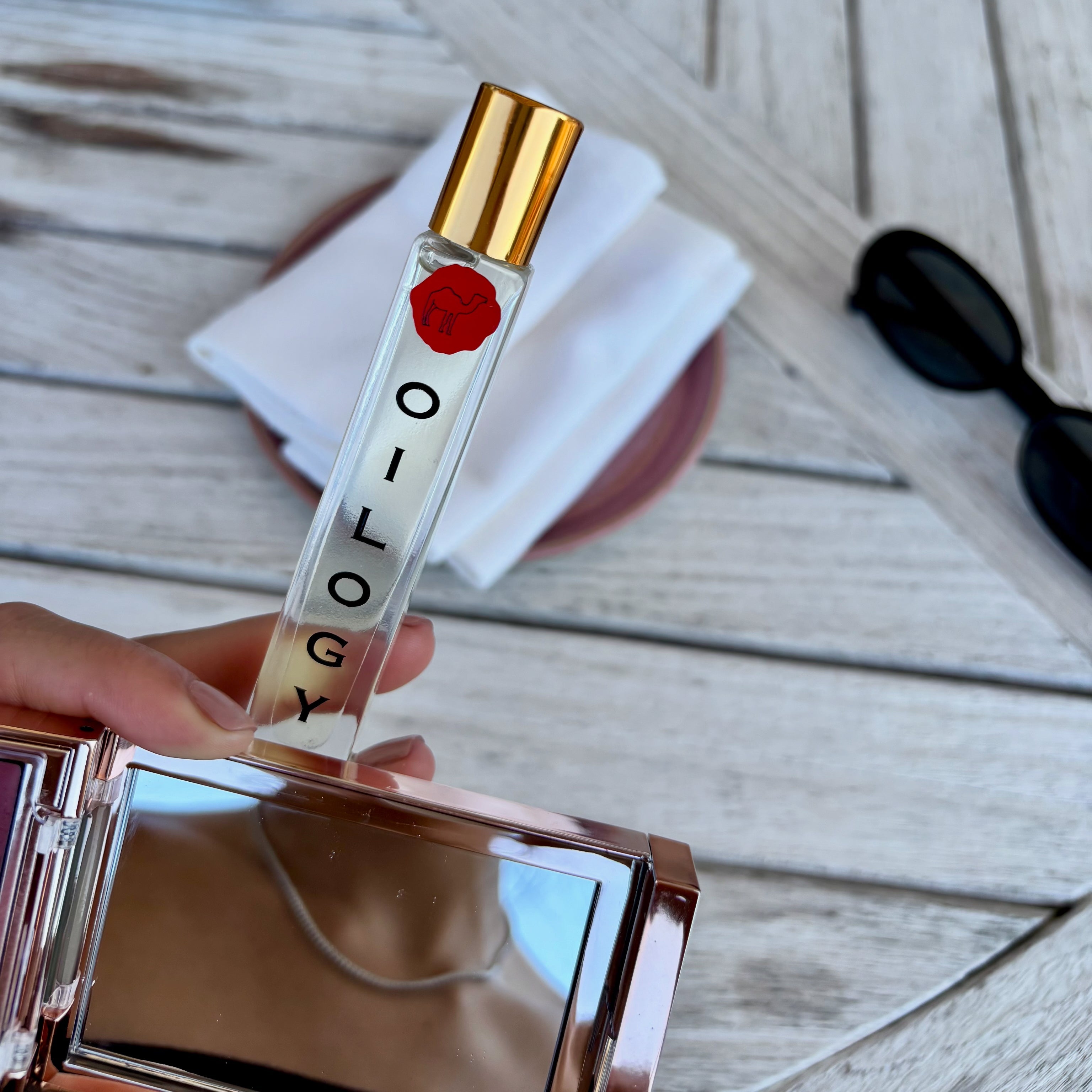 Hand holding a small bottle labeled 'Ology' with a red cap on a wooden surface.